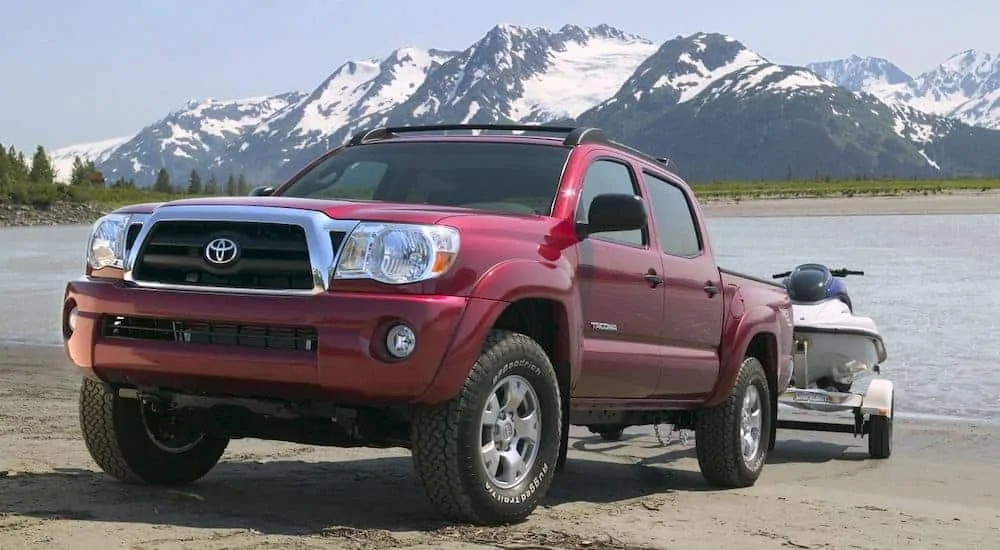 A burgundy 2007 used Toyota Tacoma is towing a jet ski in front of a lake and mountains.
