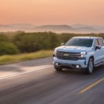 A white 2021 Chevy Silverado 1500 is driving on a rural highway at sunset.