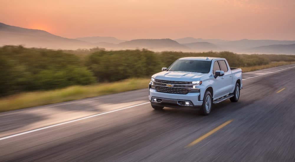 A white 2021 Chevy Silverado 1500 is driving on a rural highway at sunset.