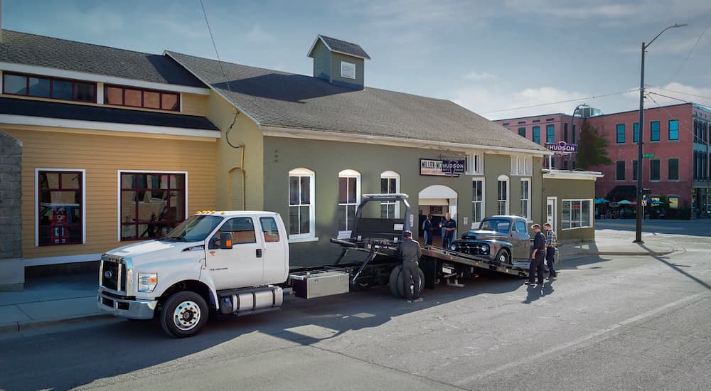 A white 2021 Ford F-650 is parked while a classic truck is being loaded on the bed.