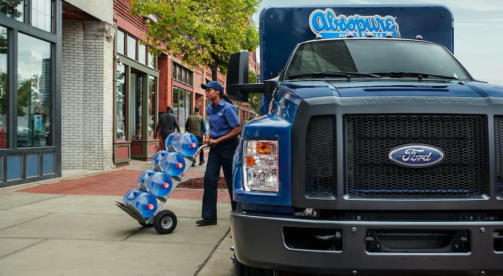 A blue 2021 Ford F-750 is shown parked on the street with a worker delivering water jugs.