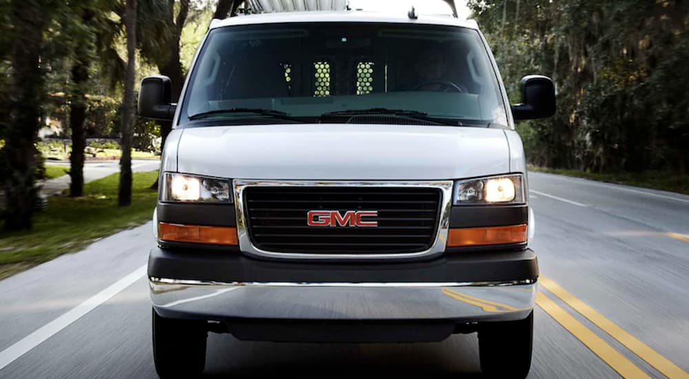 A white 2021 GMC Work Van, Savana, is shown from the front driving on a road with trees.