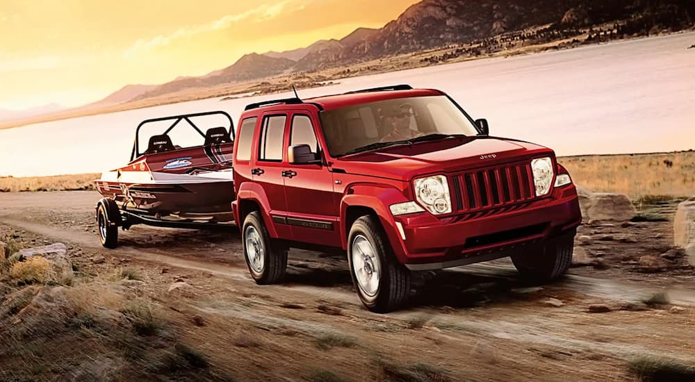 A red 2011 Jeep Liberty is towing a boat past a lake at sunset.