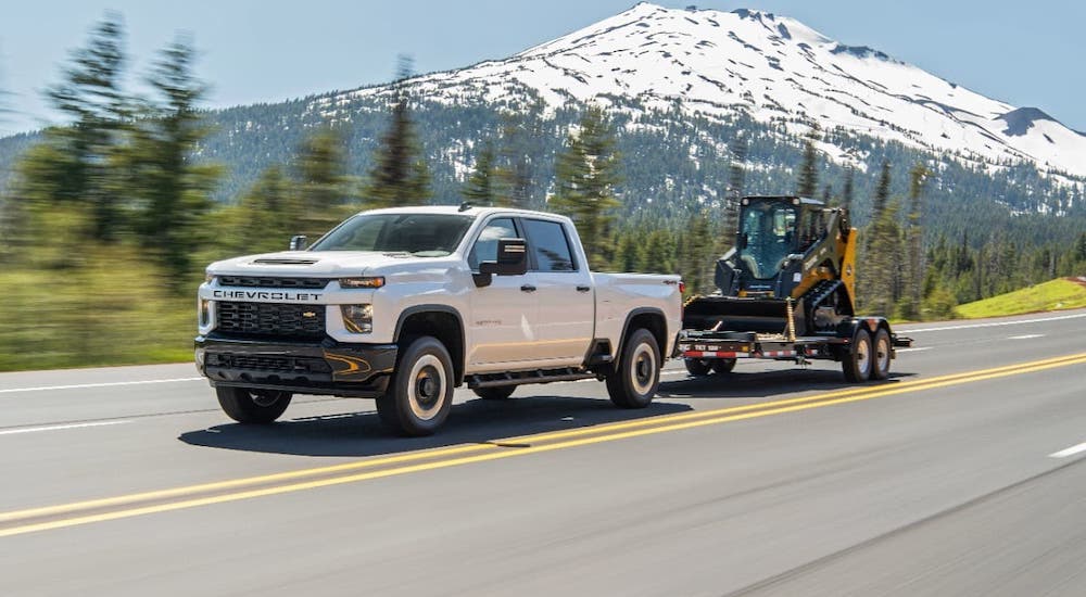A white 2021 Chevy Silverado HD is towing construction equipment on a highway in front of snow-covered mountains.