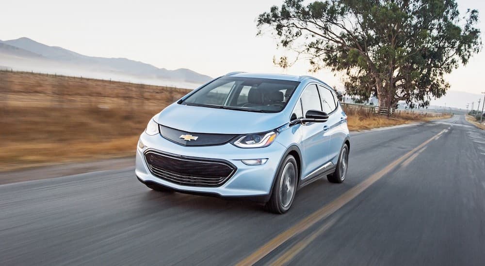 A light blue 2017 Chevy Bolt EV is driving on a rural road past mountains.
