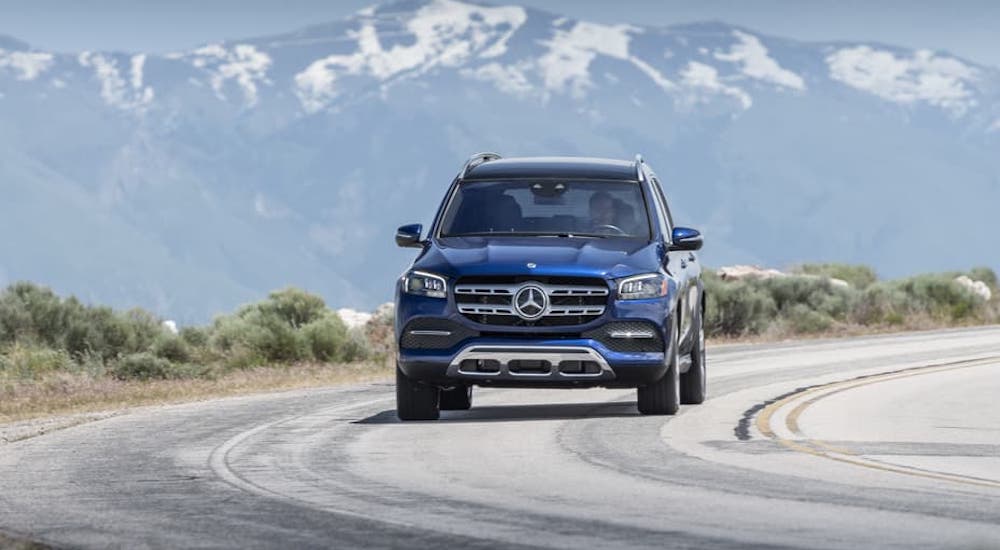 A blue 2021 Mercedes-Benz GLS SUV 450 is shown from the front on a highway in front of snow-covered mountains.