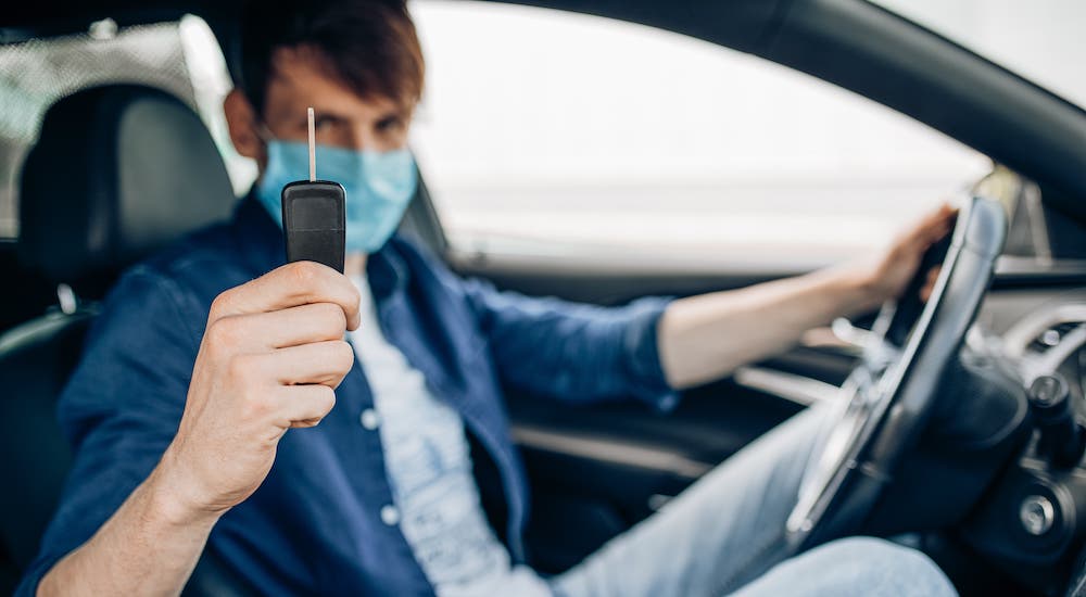 A man in a medical mask is holding a car key while sitting inside a car.