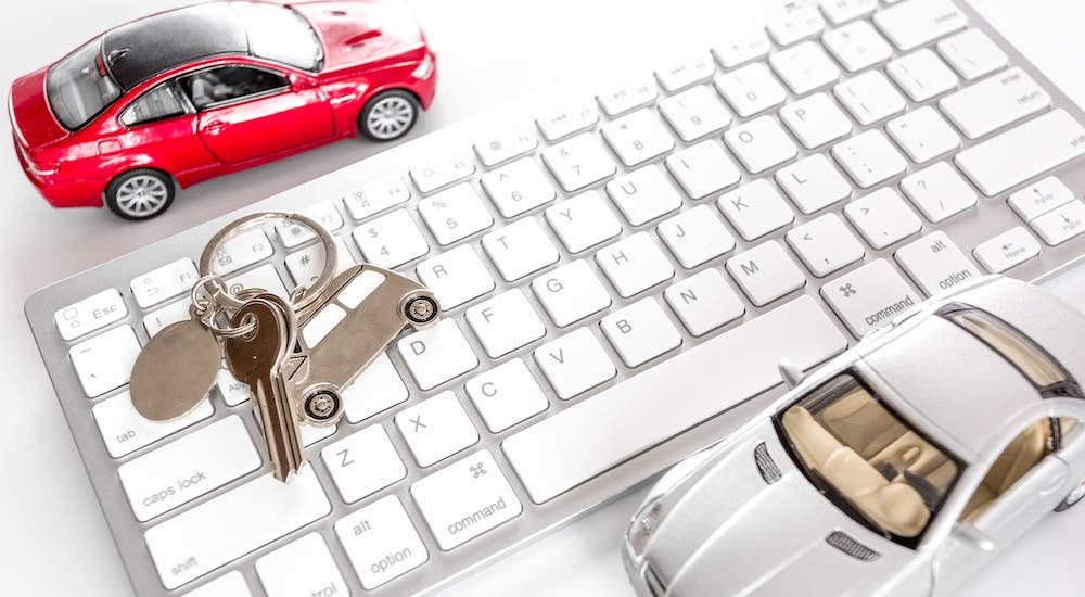 A closeup shows a red toy car, a silver toy car, and keys with a car keychain next to a computer keyboard.