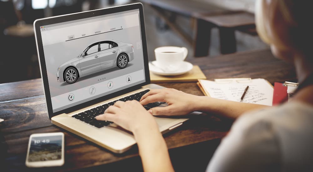 A woman is at a coffee shop and using a laptop to shop for cars at an online car dealer.