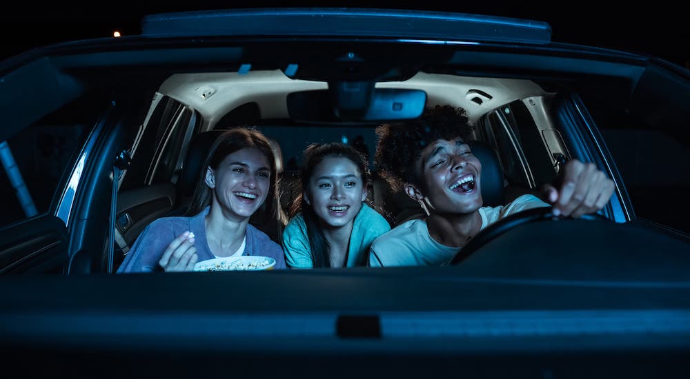 A group of teens are shown in a car at a drive-in theater at night.
