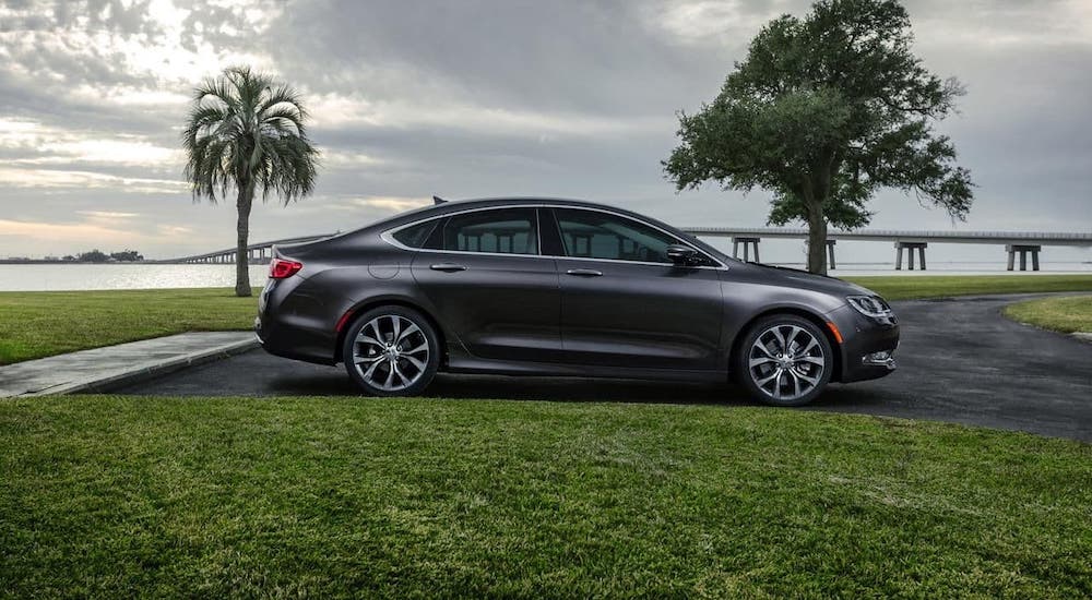 A gray 2015 Chrysler 200 is shown from the side parked in front of trees and a bay.
