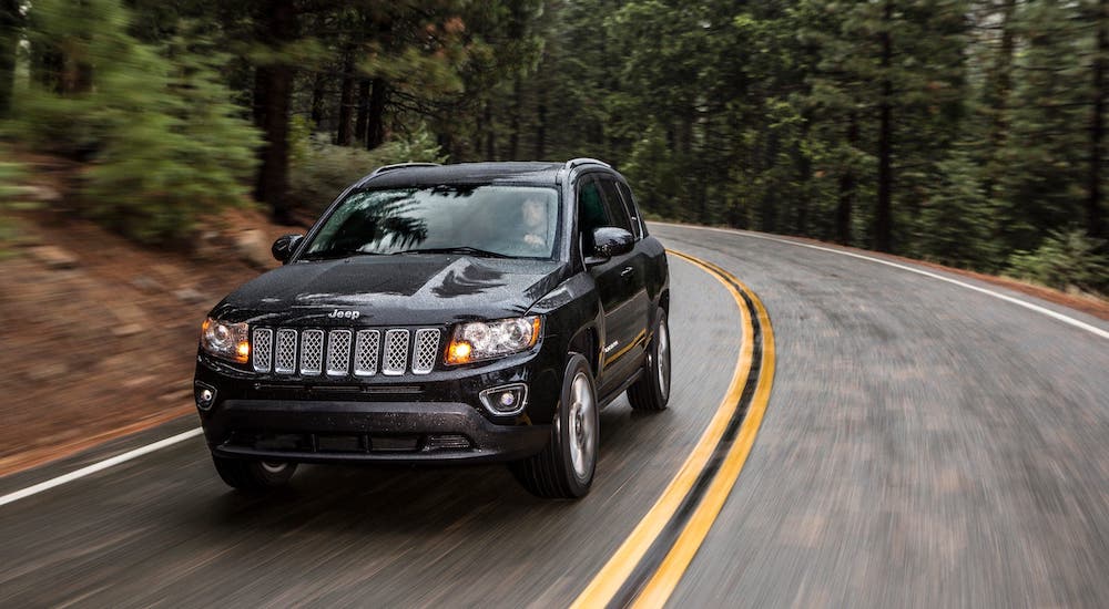 A popular used Jeep for sale, a black 2015 Jeep Compass, is driving on a woodland road in the rain.