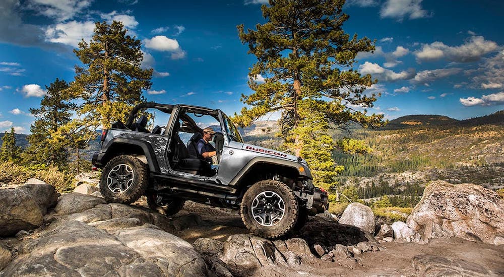A silver 2015 Jeep Wrangler Rubicon with no doors or roof is shown from the side climbing over rocks.