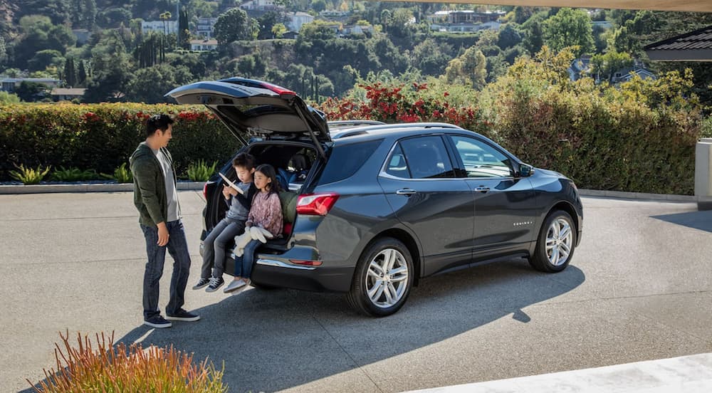 A father is next to his kids sitting in the open lift gate of a grey 2021 Chevy Equinox vs 2021 Jeep Compass in a driveway with a view.