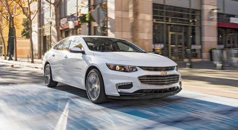 A white 2021 Chevy Malibu is driving through a city intersection.