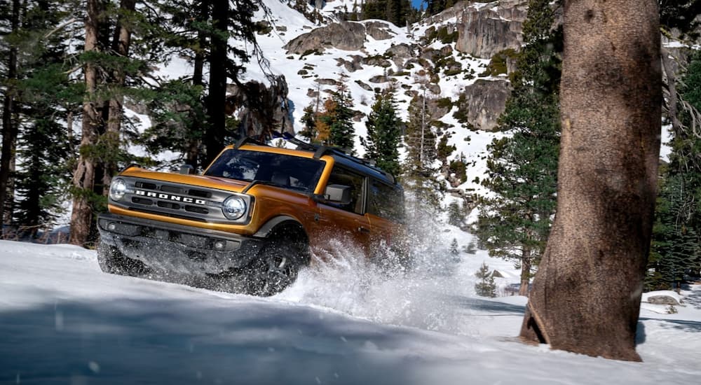 An orange 2021 Ford Bronco 2 door is off-roading in the snow in the woods.