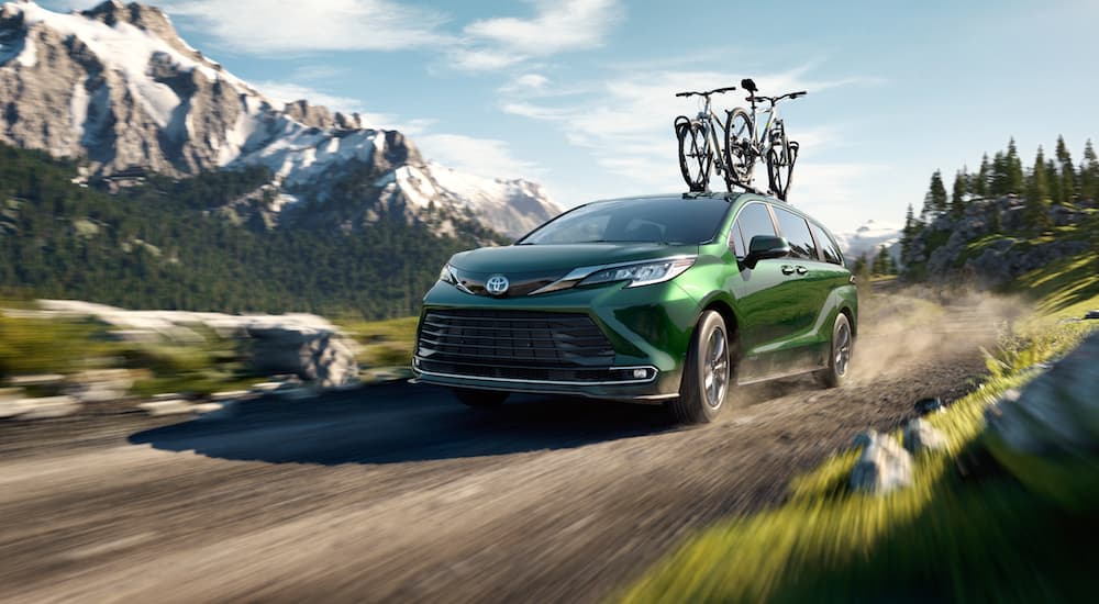A green 2021 Toyota Sienna with bikes on the roof is driving on a dirt road past mountains.