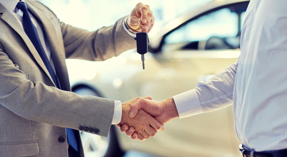 A salesman is handing a key to a buyer and shaking his hand in front of a white car.