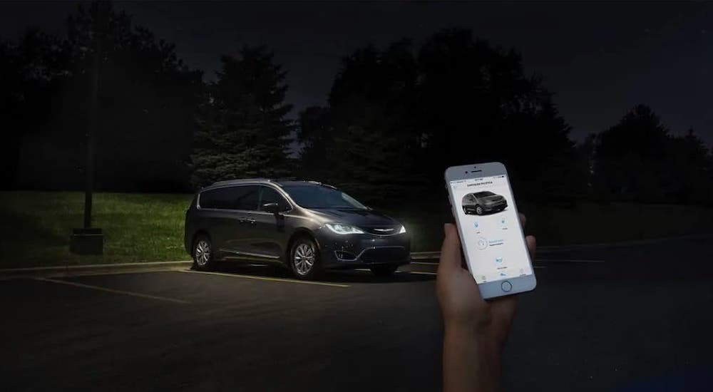 A hand is holding a phone at night and commanding the black Chrysler Pacifica in the background.