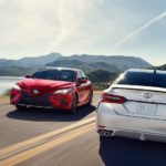 A red and a white 2018 Toyota Camry are passing each other on a mountain road next to a lake.
