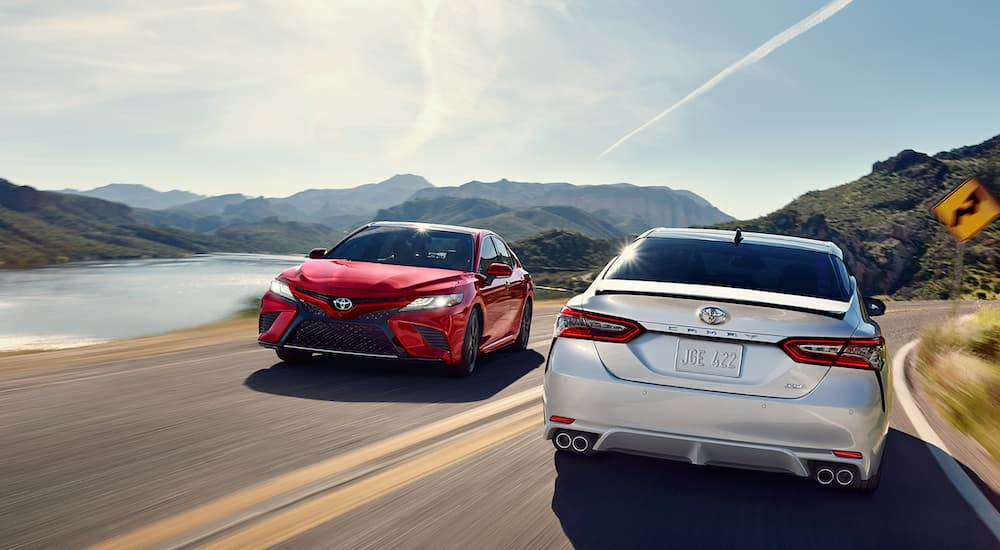 A red and a white 2018 Toyota Camry are passing each other on a mountain road next to a lake.