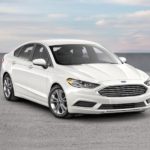 A white 2020 Ford Fusion is parked in an empty lot against a cloudy sky.