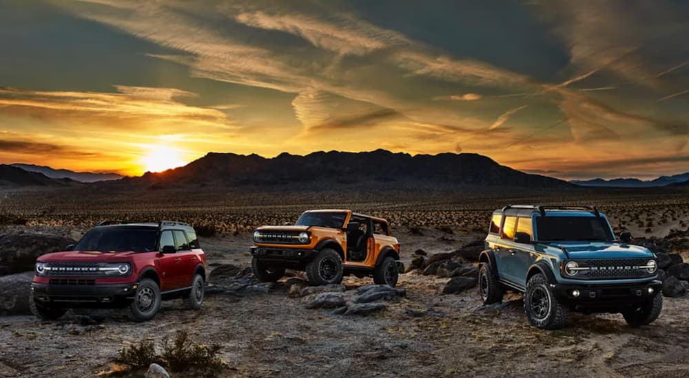 A red 2021 Ford Bronco Sport, a yellow 2021 Ford Bronco 2-door, and a blue 2021 Ford Bronco 4-door are parked in the desert at sunset.