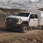 A white 2021 Ford Chassis Cab F-450 with toolbox is driving in front of a dirt pile.