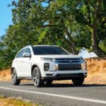 A white 2021 Mitsubishi Outlander Sport is driving on a rural highway in front of trees.