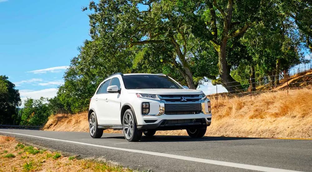 A white 2021 Mitsubishi Outlander Sport is driving on a rural highway in front of trees.
