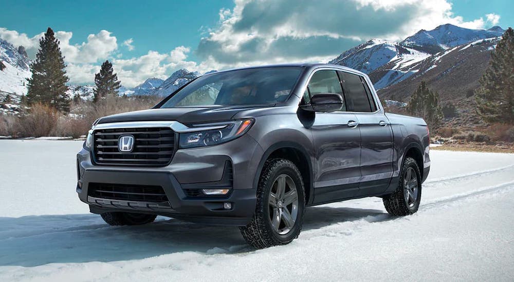 A gray 2021 Honda Ridgeline is driving in the snow in front of evergreen trees and snow-covered mountains.