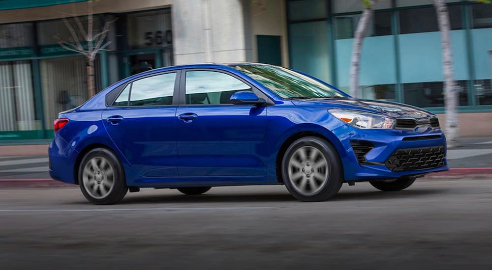 A blue 2021 Kia Rio sedan is driving in front of an office building.