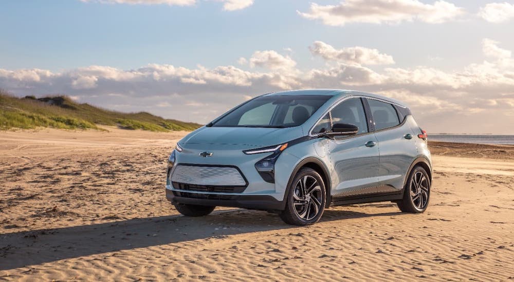 A new model of a classic Chevy EV, a pale blue 2022 Chevy Bolt EV, is parked on a beach.