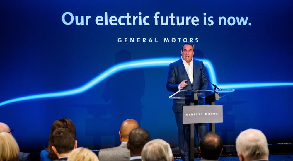 Mark Reuss, the president of GM, is speaking at a podium in front of a blue screen that reads 'our electric future is now.'