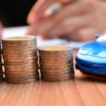 A blue die-cast car is on a wooden table next to three stacks of coins.