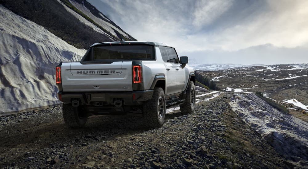 A white 2022 GMC Hummer EV from a GMC dealer is shown from the rear driving on a dirt trail next to snow-covered mountains.