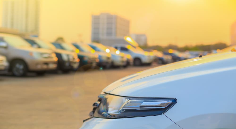 A close up shows the headlight of a car with a blurred row of cars in the background.