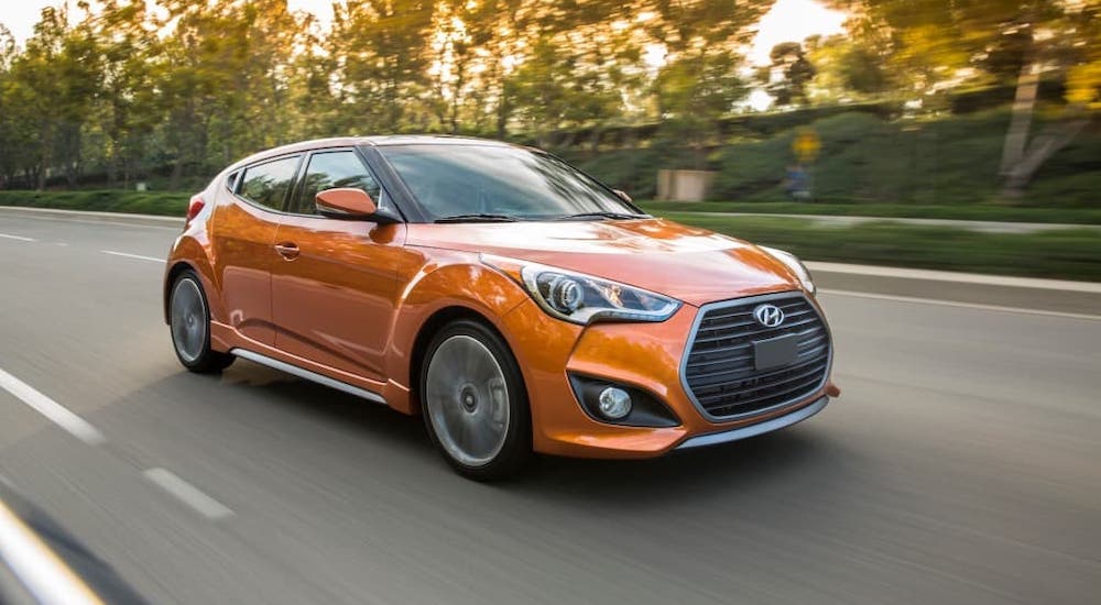 An orange 2017 Hyundai Veloster is driving on a street in front of trees.