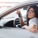 A smiling young woman is holding up car keys inside a white sedan at a used car dealership.