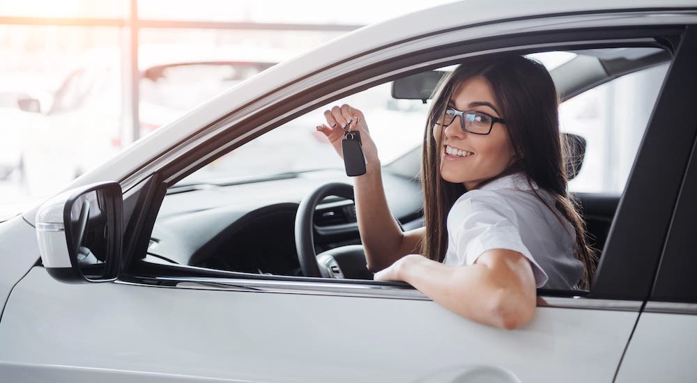 A smiling young woman is holding up car keys inside a white sedan at a used car dealership.