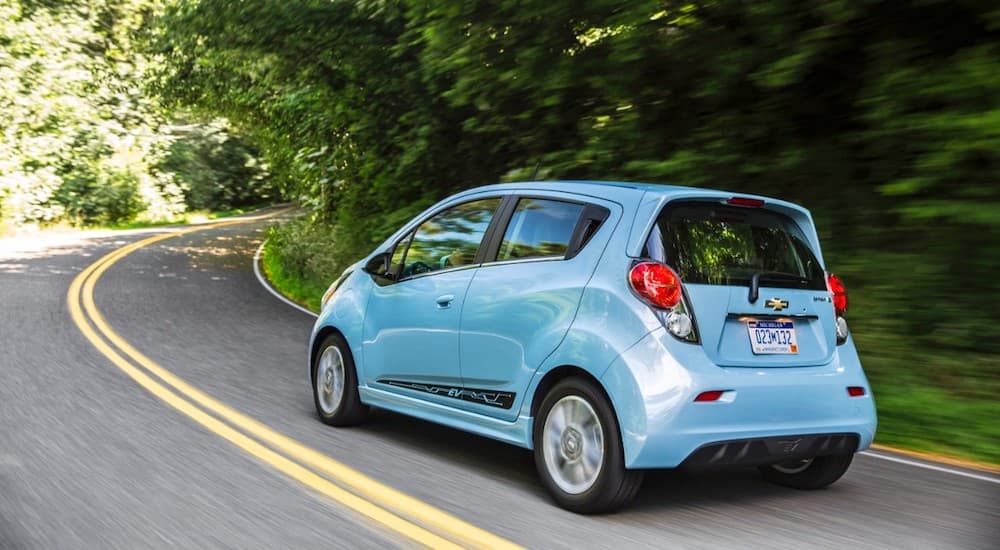A blue 2015 Chevy Spark EV is driving around a corner past trees.