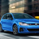 A blue 2021 Volkswagen Golf GTI is shown from the front driving through a city.