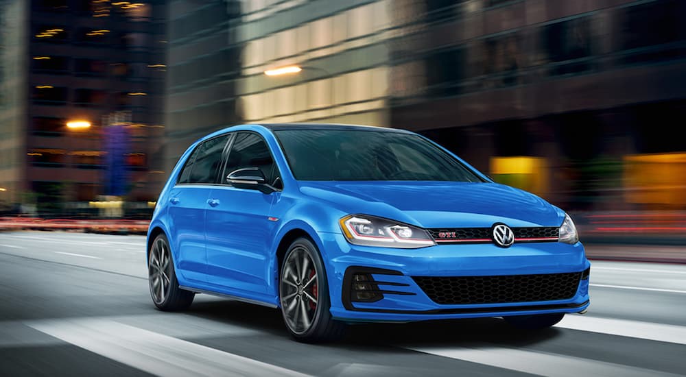 A blue 2021 Volkswagen Golf GTI is shown from the front driving through a city.