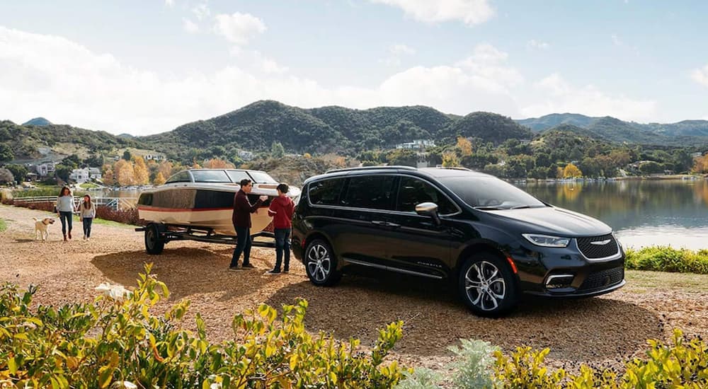 A black 2021 Chrysler Pacifica Pinnacle is towing a boat on a dirt road in front of a lake.