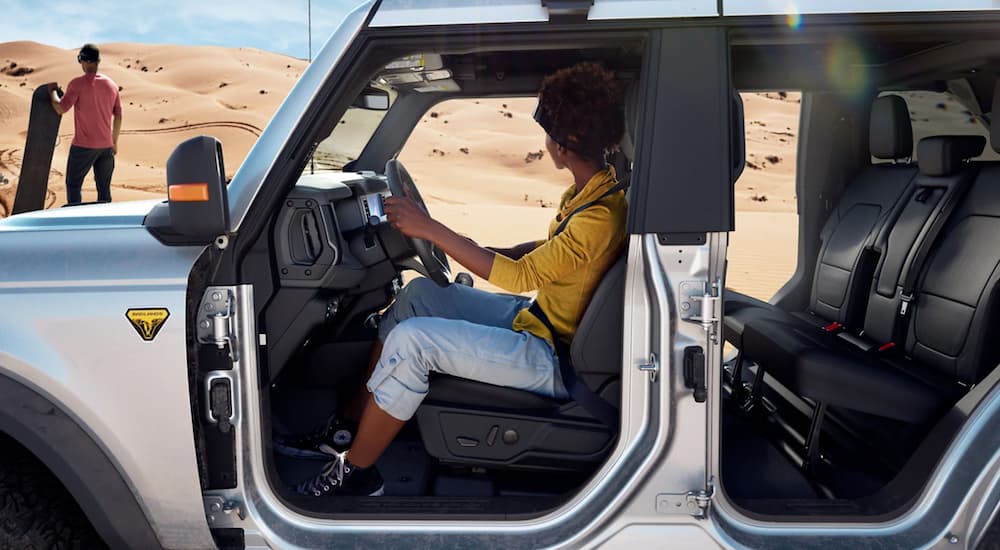 A silver 2021 Ford Bronco 4-door is shown from the side with no doors and a woman in the driver's seat.