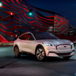 A white 2021 Ford Mustang Mach-E drives through an urban street at night.