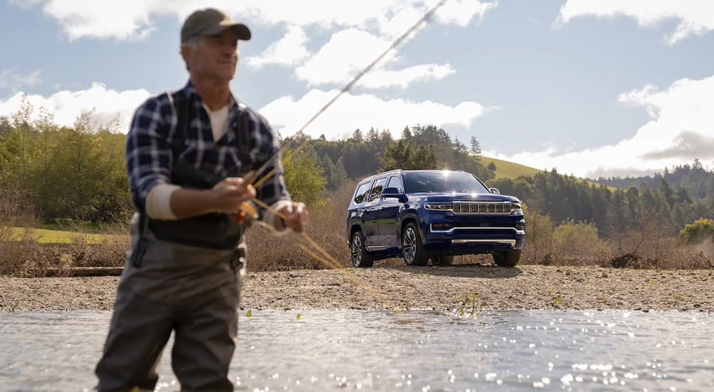 A blue 2022 Grand Wagoneer is parked on the edge of the river, a man is fishing in the river.