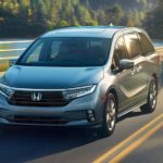 A gray 2022 Honda Odyssey is driving in front of a river and trees.