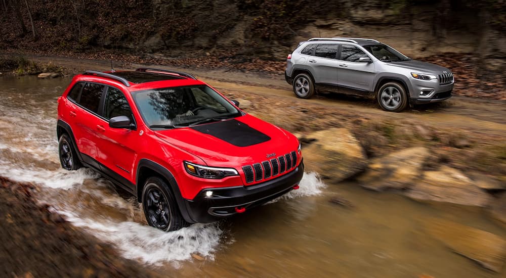 A red 2019 Certified Pre-Owned Jeep Cherokee is off-roading in a river next to a silver 2019 Cherokee on a dirt road.