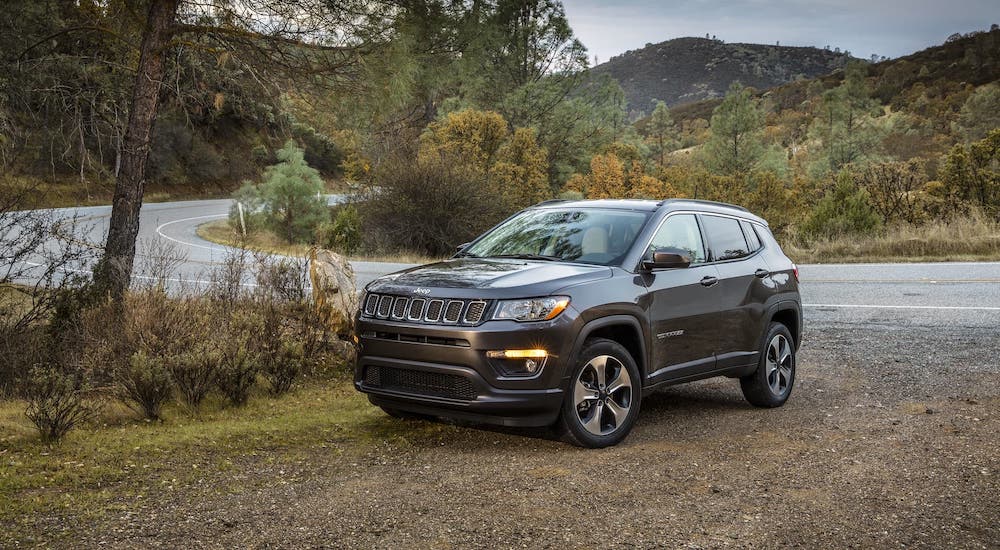 A gray 2020 Used Jeep Compass is parked on a dirt area next to a highway.